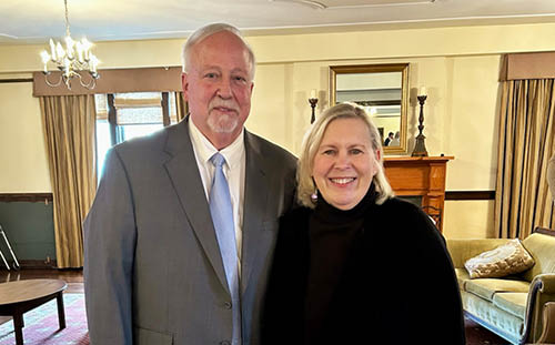 A man in a gray suit and a woman in black stand smiling, feeling it’s a privilege to serve, in a warmly lit living room with vintage decor.