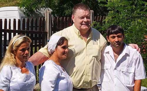 Four adults, part of Romania mission trips, stand together outdoors in front of a wooden fence and greenery.