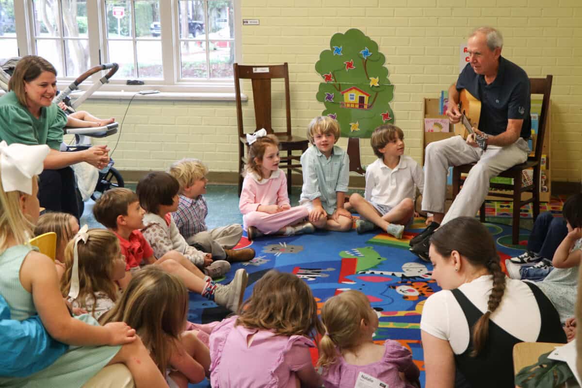 An adult plays guitar for children seated on a colorful rug at First Presbyterian Baton Rouge, with two adults nearby.