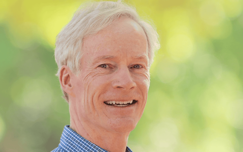 Older man with light hair smiling, wearing a blue checked shirt, outdoors near First Presbyterian Baton Rouge’s vibrant green and yellow backdrop.