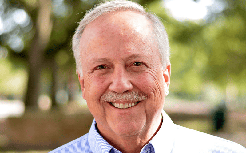 An older man with gray hair smiles outdoors in sunlight, wearing a light blue shirt—representing First Presbyterian Baton Rouge.
