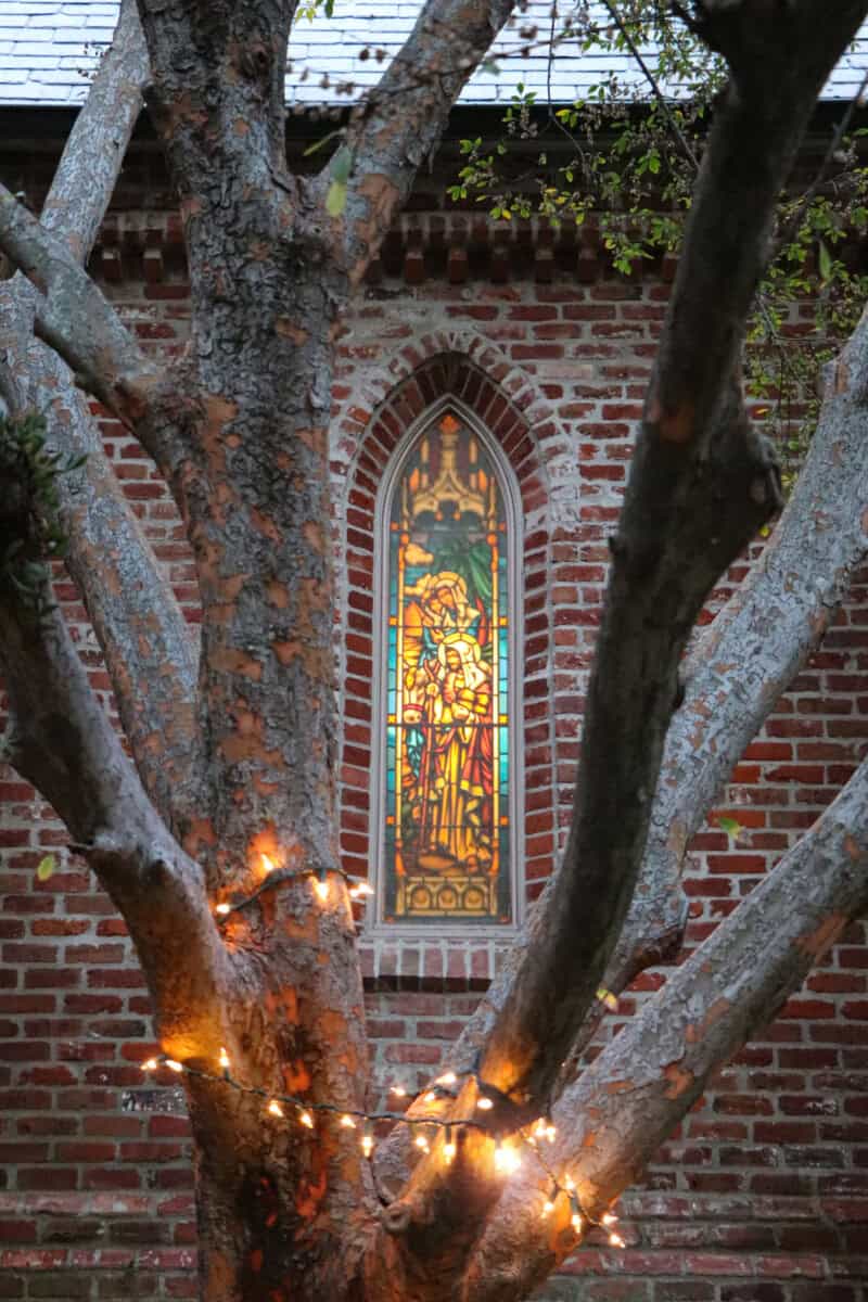 A tree with string lights stands in front of a brick building with a stained glass window depicting a religious figure.
