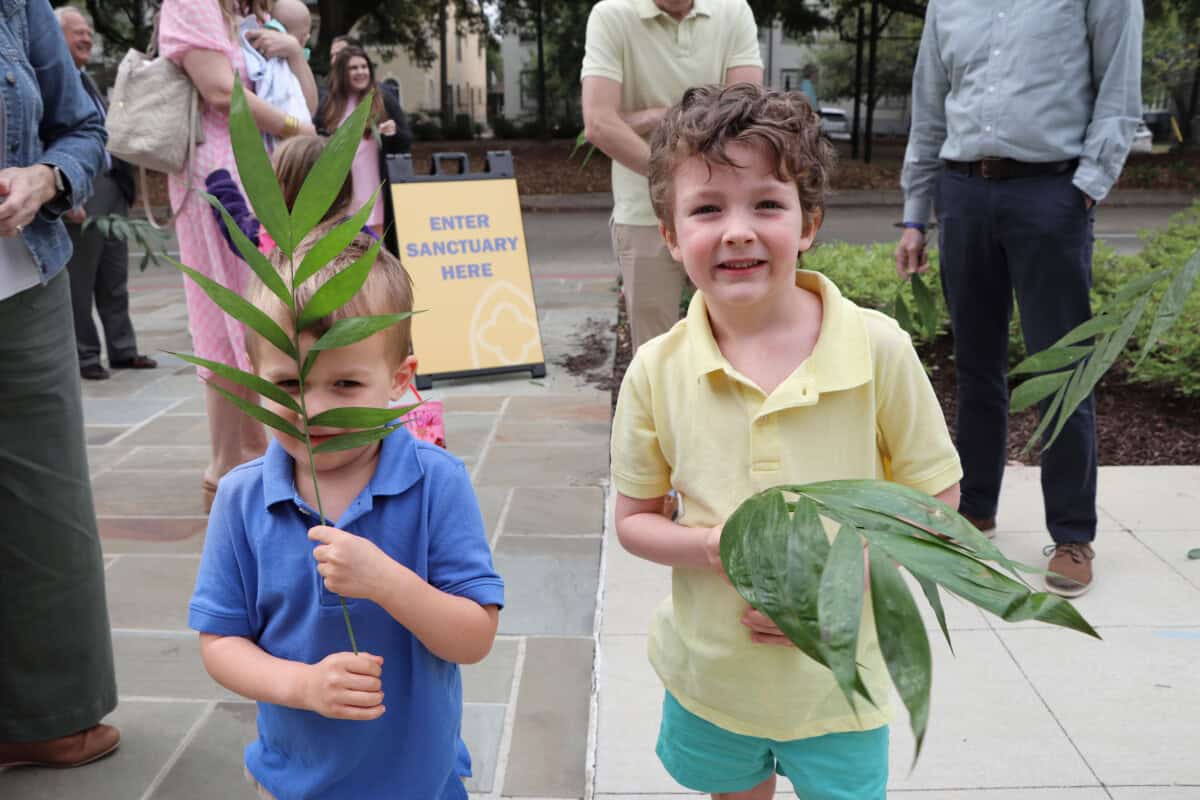 Two young boys holding leafy branches stand outside near adults; one boy hides his face with the branch. A sign reads 