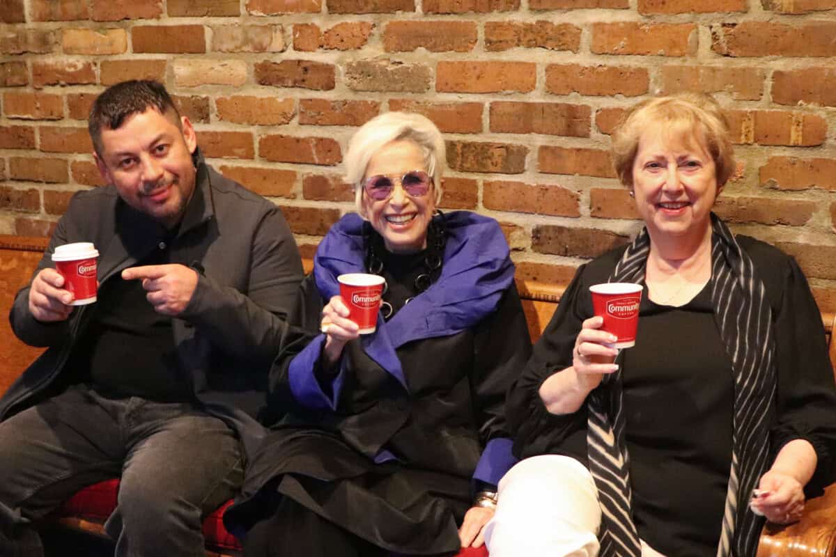 Three people sit on a bench against a brick wall at First Presbyterian Baton Rouge, each holding a red coffee cup and smiling at the camera.