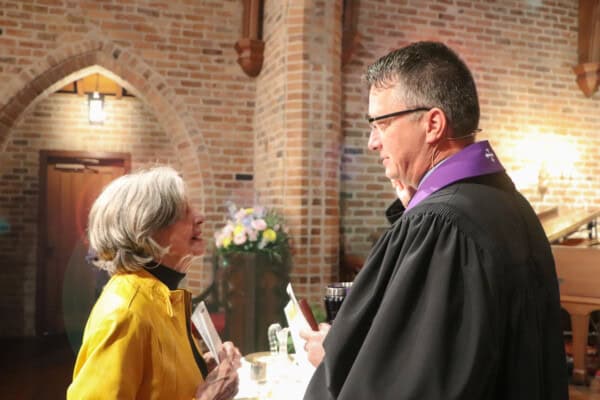 On Sunday mornings, an older woman in a yellow jacket talks to a man in a black robe inside a church with brick walls and flowers.