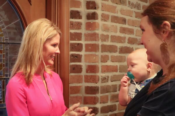 Two women stand by a brick wall, one holding a baby with a pacifier—just what to expect on Sunday mornings, sharing smiles and conversation.