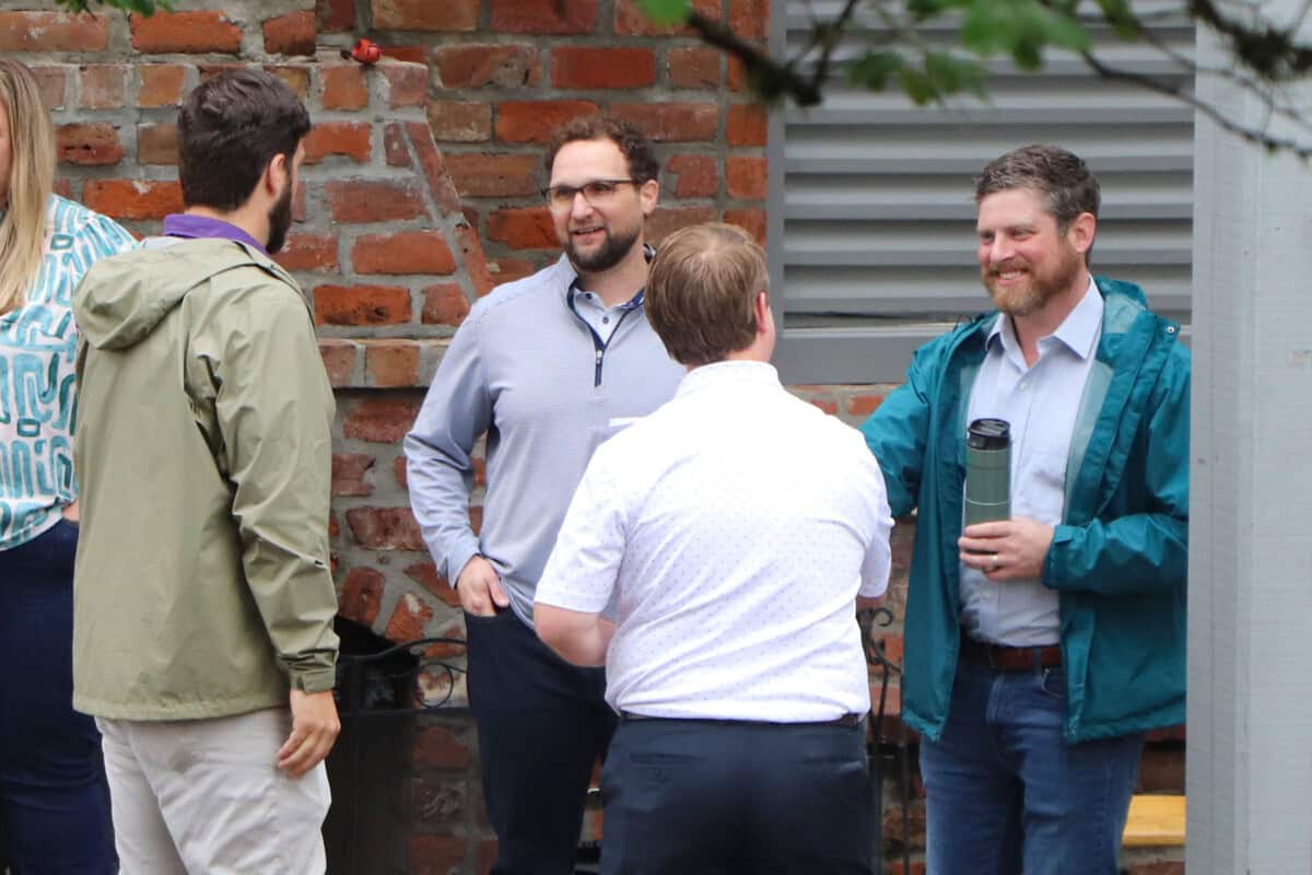 Four men are standing outside near a brick wall, smiling and chatting on a Sunday morning; one holds a travel mug.
