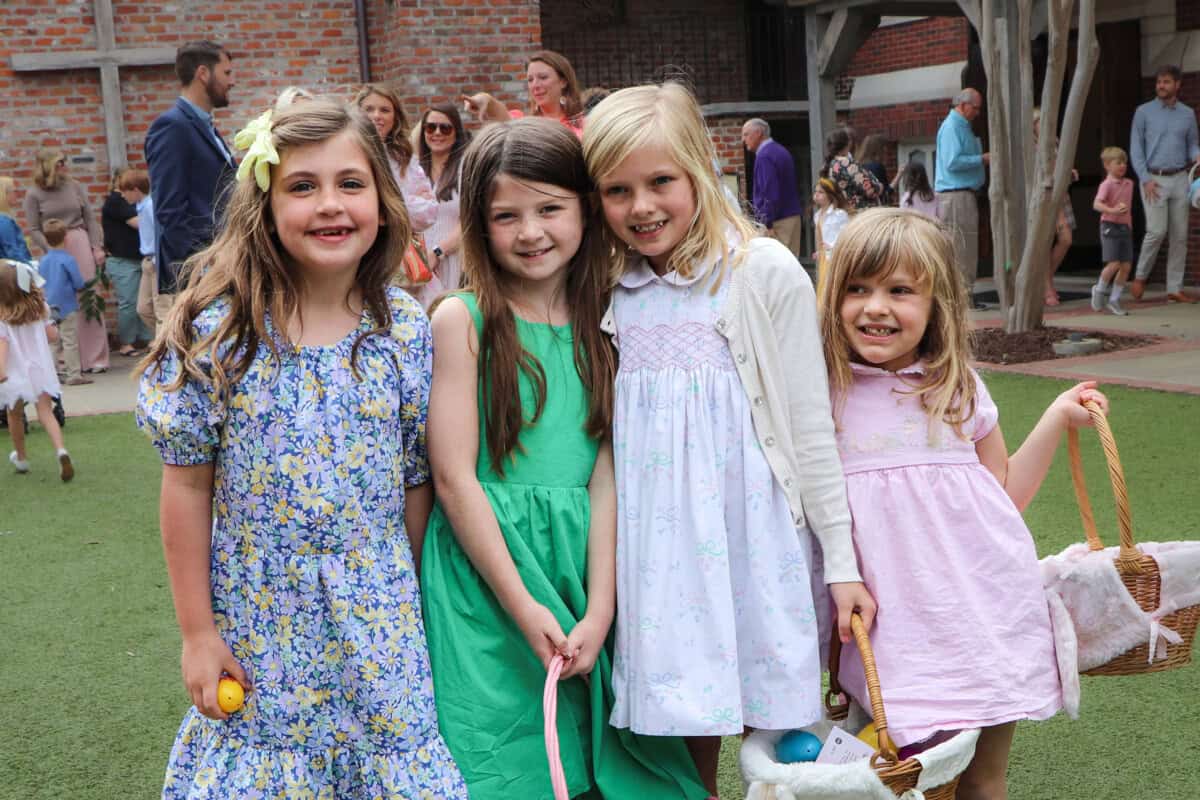 Four young girls stand together outside on grass, smiling at the camera. They wear spring dresses and hold Easter baskets and eggs.