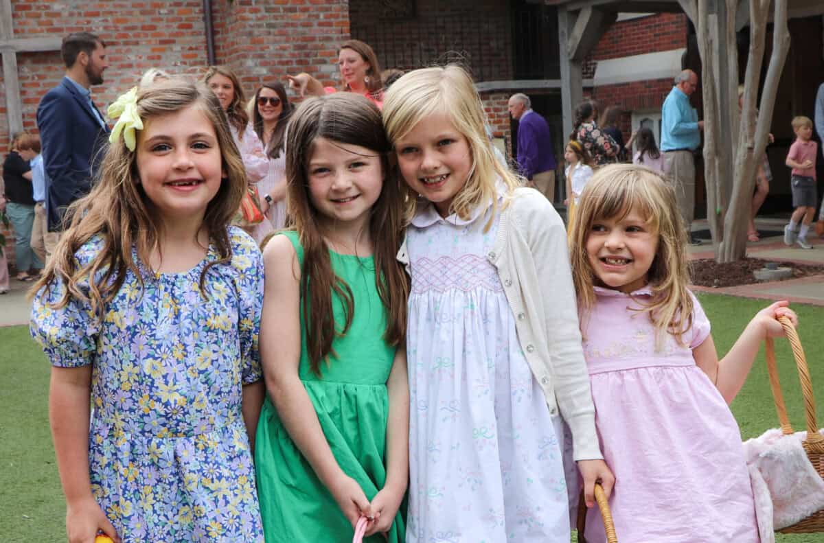 Four young girls stand together outside First Presbyterian Baton Rouge, smiling in colorful spring dresses with people in the background.