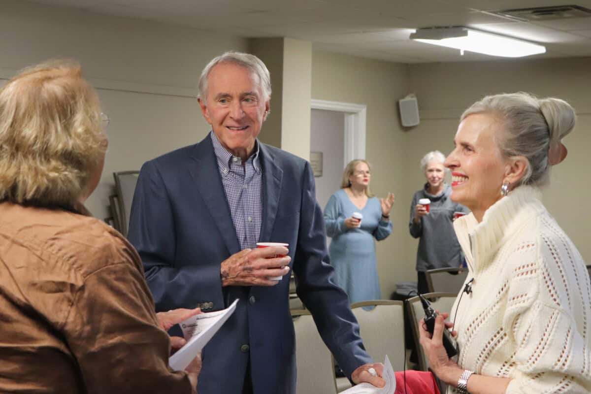 Three people smile and chat at a gathering, holding papers and cups at First Presbyterian Baton Rouge; others mingle in the background.