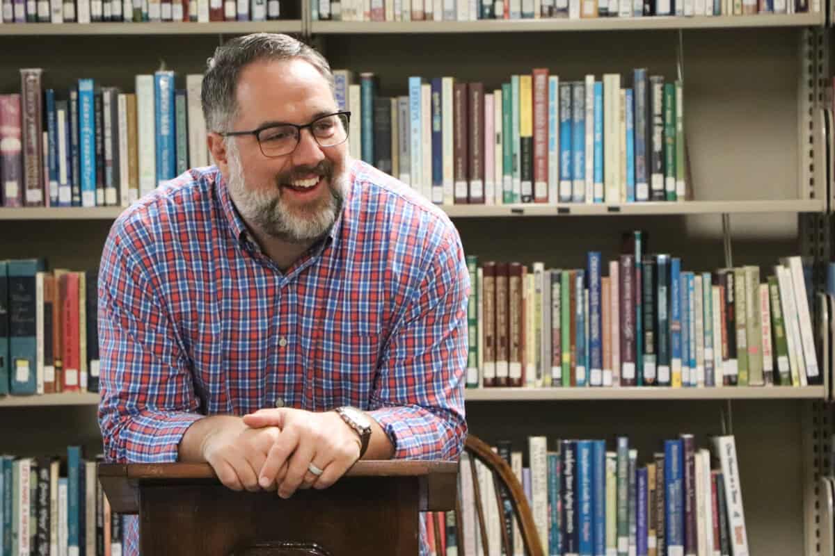 A man with glasses and a beard, in plaid shirt, smiles at a podium in the library of First Presbyterian Baton Rouge.