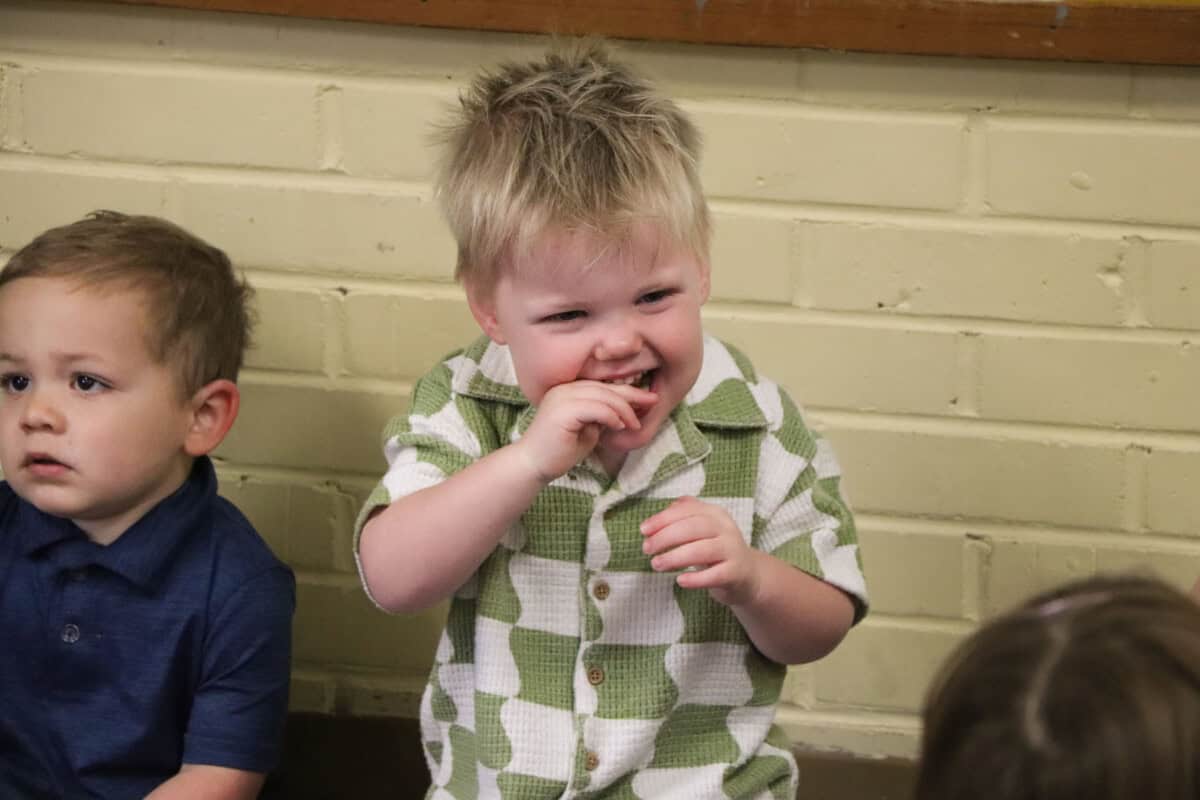 Two young children sit indoors against a yellow brick wall at First Presbyterian Baton Rouge; one touches his mouth in a green and white shirt.
