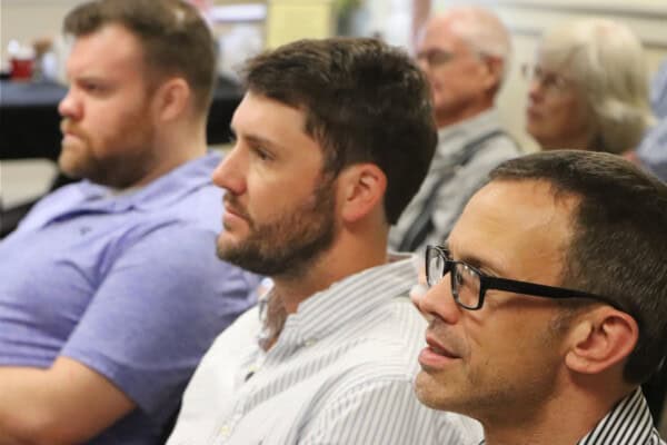 Three men seated in a row at a First Presbyterian Baton Rouge event, with others blurred in the background at a church meeting.
