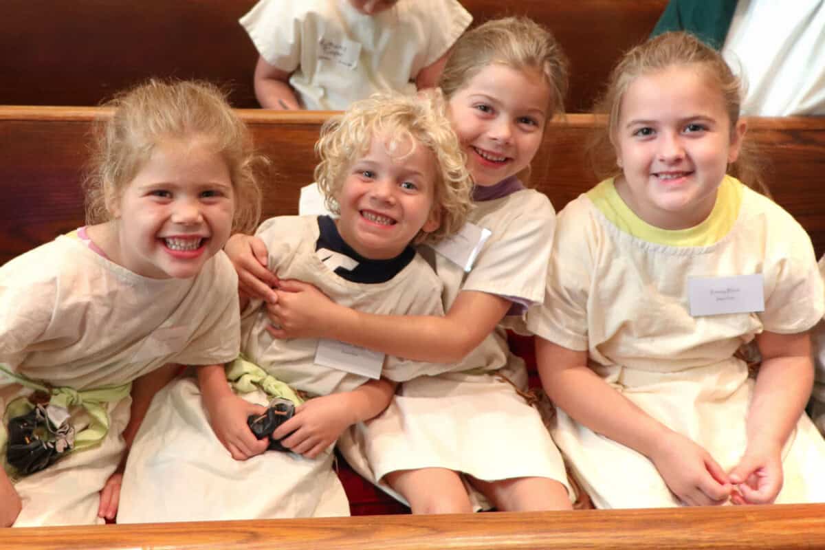 Four young children in light tunics sit closely on a wooden bench, smiling—just a glimpse of Sunday Mornings at What to Expect’s Sunday Service.