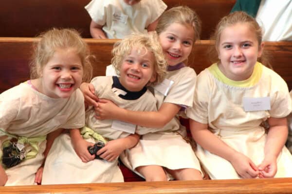 Four young children in light tunics sit closely on a wooden bench, smiling—just a glimpse of Sunday Mornings at What to Expect’s Sunday Service.