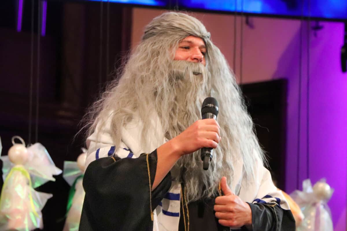 A person in a long gray wig and costume gives a thumbs up on stage with purple lighting—think Sunday mornings with a twist.