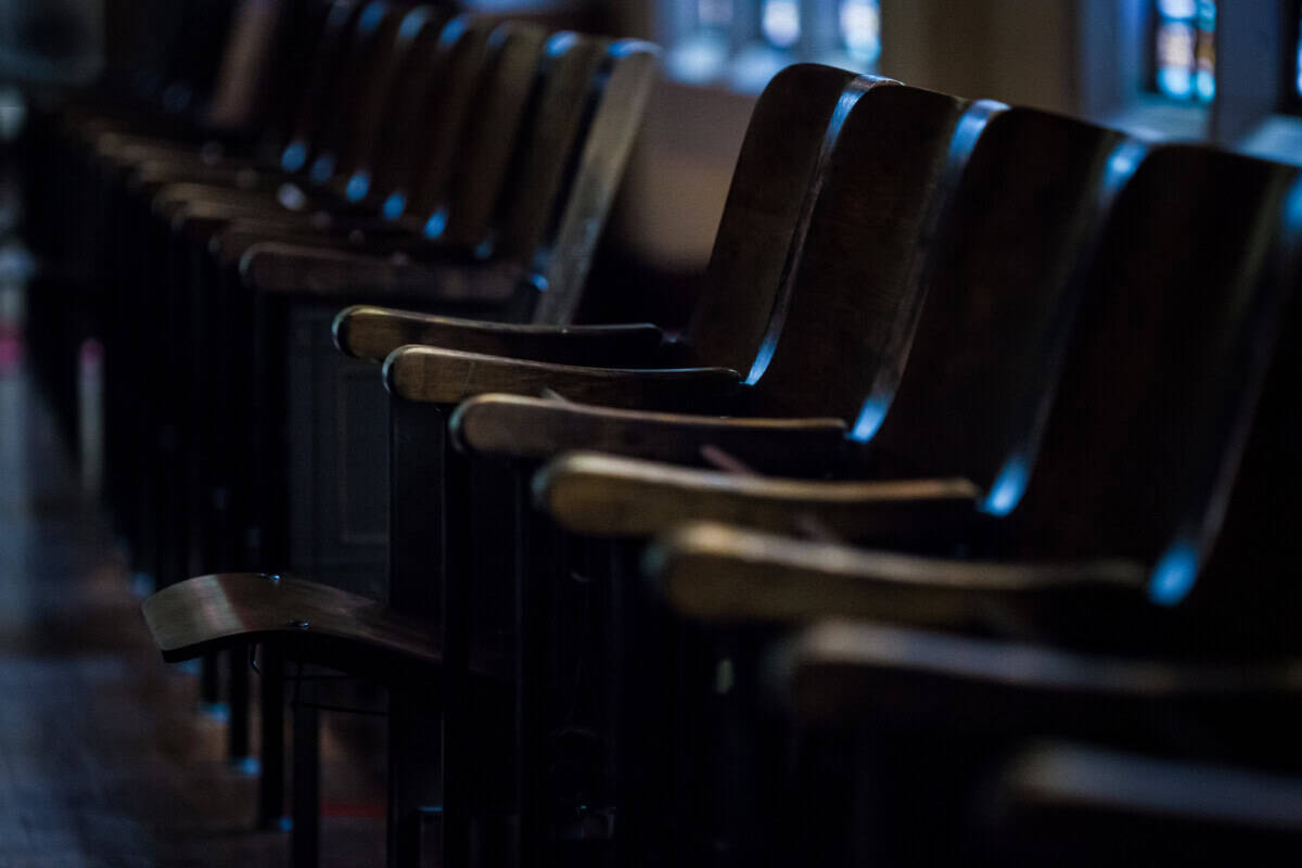 A row of empty, dark wooden seats in an auditorium or theater, lit by dim light from windows in the background.