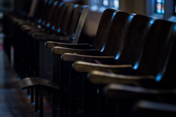 A row of empty, dark wooden seats in an auditorium or theater, lit by dim light from windows in the background.