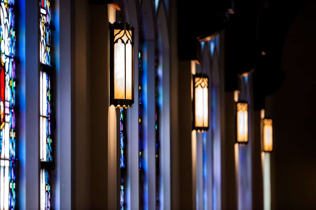 A row of lit wall lanterns glows beside arched blue stained glass at First Presbyterian Baton Rouge’s dimly lit interior.
