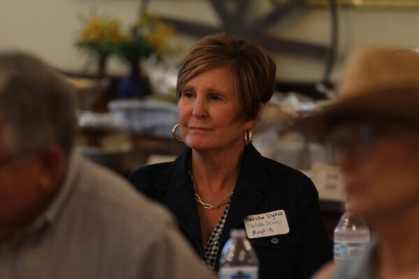 A woman with short hair and hoop earrings sits at a table at First Presbyterian Baton Rouge, wearing a name tag and dark jacket, water bottles in front.
