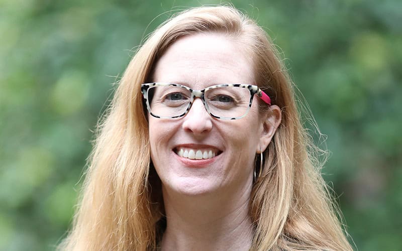 A woman with long light brown hair and patterned glasses, one of our Deacons, smiles outdoors in front of a blurred green background.