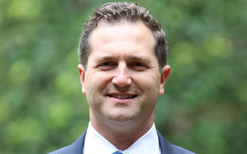A man in a suit and tie, possibly one of the deacons, smiles at the camera with greenery blurred in the background.