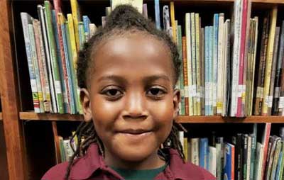 A young child with long braided hair smiles, standing before a bookshelf at Baton Rouge Presbyterian Church.