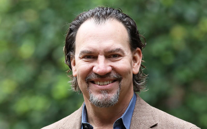 A middle-aged elder with slicked-back hair and a goatee, wearing a brown blazer, smiles in front of a blurred green background.