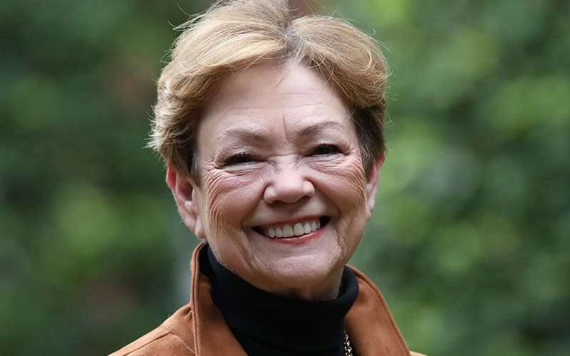 An older woman, possibly one of the elders, smiles outdoors in a brown jacket and black turtleneck, framed by lush green foliage.