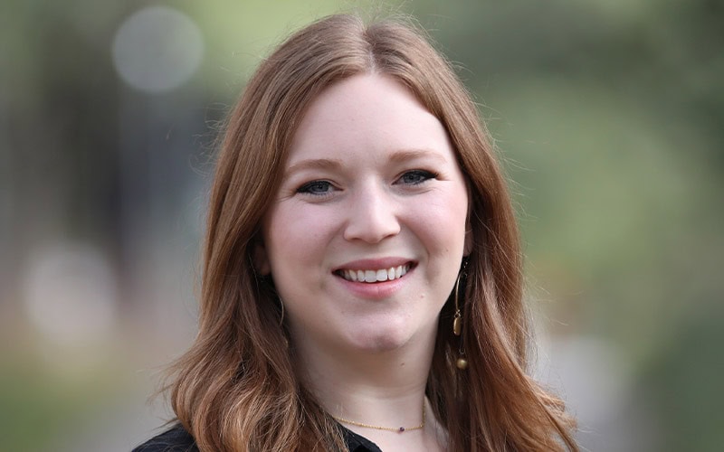 A woman with long, wavy, light brown hair—one of the deacons—smiles at the camera outdoors in a black top and gold earrings.