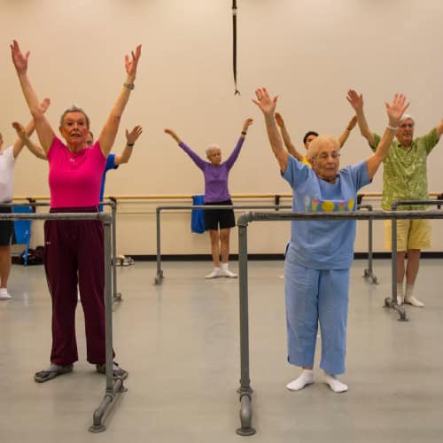 Older adults enjoy a fitness class in a studio at First Presbyterian Baton Rouge, standing behind barres with arms raised.