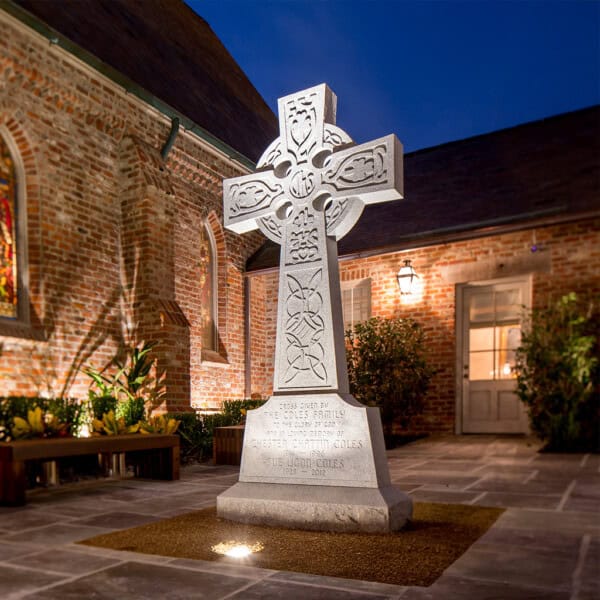 A large stone Celtic cross monument stands in a lit courtyard beside First Presbyterian Baton Rouge at night.