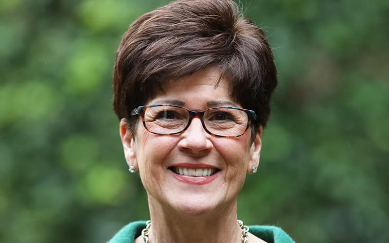 A smiling woman with short brown hair and glasses, possibly among the deacons, stands outdoors in green, framed by blurred foliage.