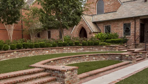 Curved brick seating and steps in an outdoor amphitheater at First Presbyterian Baton Rouge, with lush landscaping and a brick building behind.