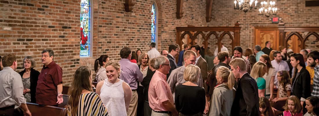 A group socializes and converses inside First Presbyterian Baton Rouge, surrounded by brick walls and stained glass windows.