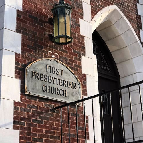 A sign reading "First Presbyterian Baton Rouge" is mounted on a red brick building next to a door and large wall lantern.