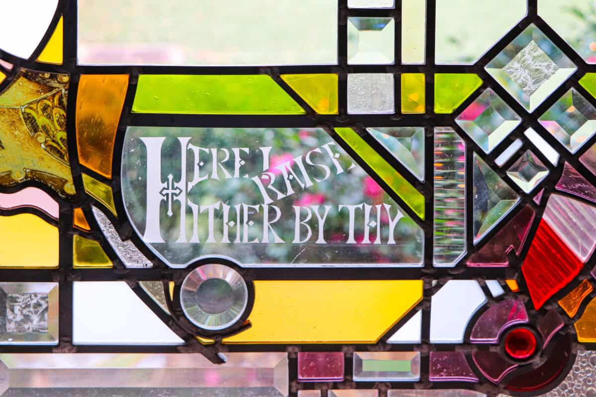 Stained glass window with the text “Here I raise mine Ebenezer; hither by thy” in white, surrounded by colorful geometric patterns.