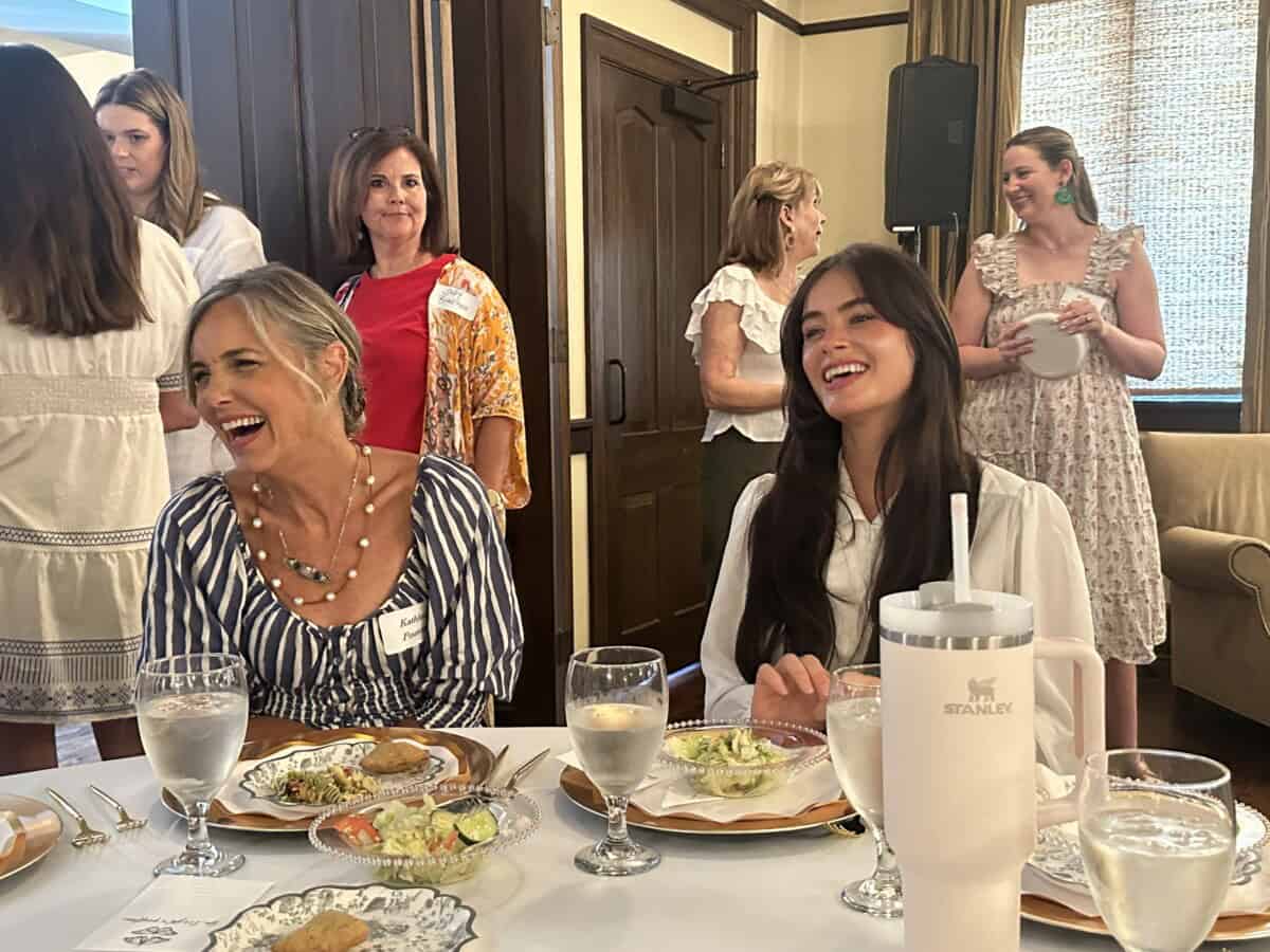 Two women sit at a table with plates of food and water glasses, smiling and laughing, while others stand and talk in the background.