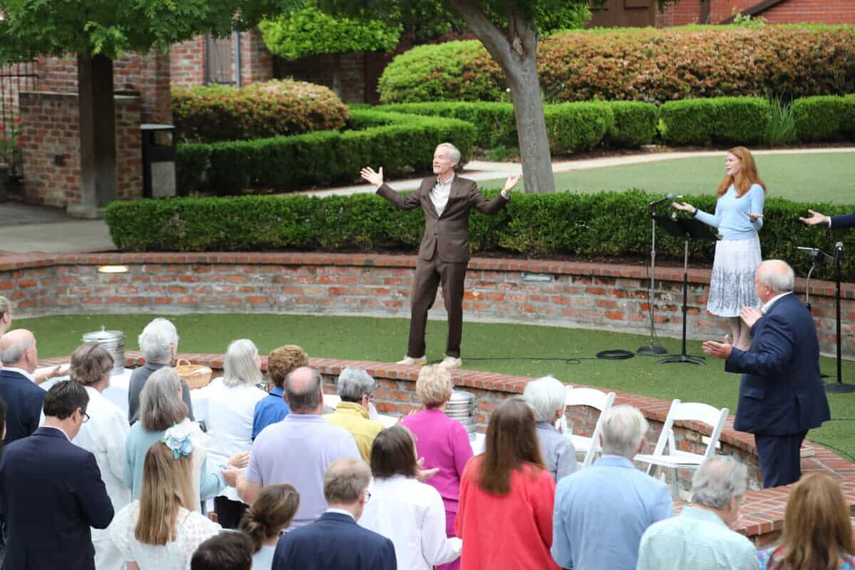 A person in a suit raises their arms on an outdoor stage at First Presbyterian Baton Rouge, as a woman stands nearby at a music stand.