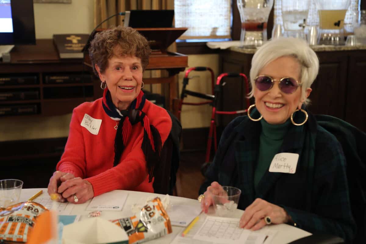 Two older women sit at a table, smiling with snacks and drinks, welcoming guests to First Presbyterian Baton Rouge.