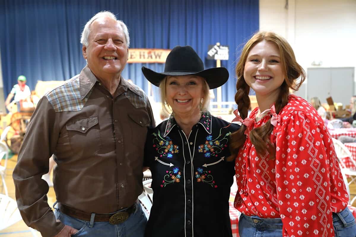 Three people in western-style clothing smile indoors at a First Presbyterian Baton Rouge event with tables and a blue curtain behind them.