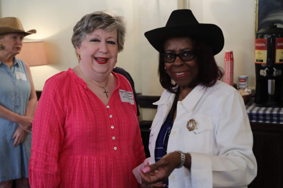 Two women smile at an indoor event at First Presbyterian Baton Rouge—one in a pink dress, the other in a white jacket and black hat.