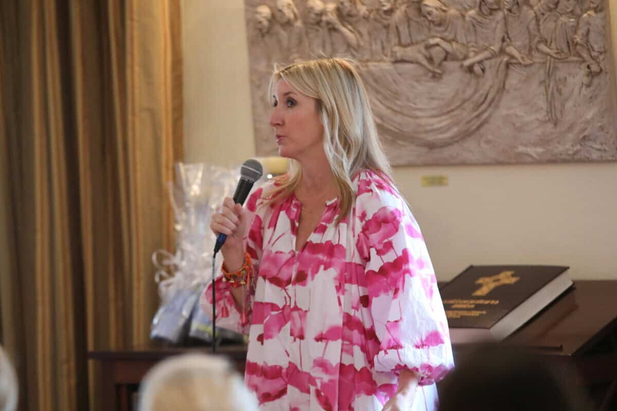 A woman in a pink and white dress speaks into a microphone in an indoor setting with people listening.