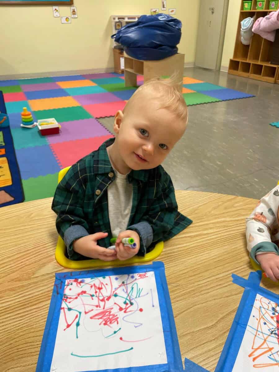A toddler sits at a table in a classroom at First Presbyterian Baton Rouge, drawing colorful scribbles on paper taped to the table.