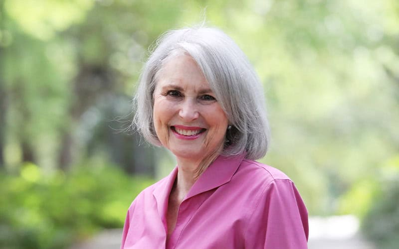An older woman with gray hair and a pink shirt smiles outdoors, embodying the warmth and wisdom of elders among lush greenery.