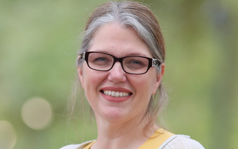 A woman with glasses and gray hair pulled back is smiling outdoors, embodying the warmth of elders against a blurred green background.