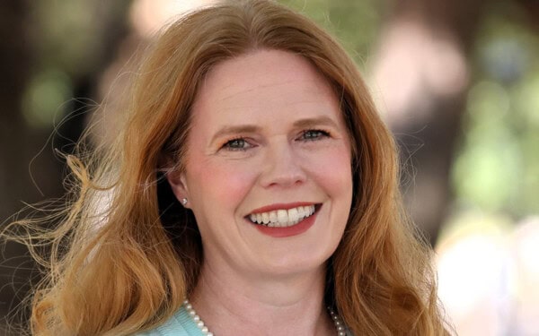 A woman with long red hair smiles outdoors near trees, dressed in a blue top and pearl necklace after First Presbyterian Baton Rouge services.