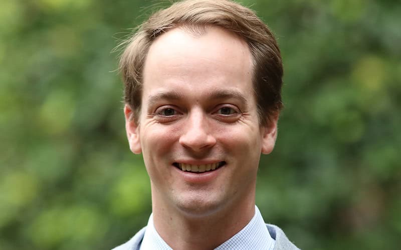 A man with short brown hair, a Deacon, wearing a suit jacket and shirt, smiling outdoors with greenery in the background.
