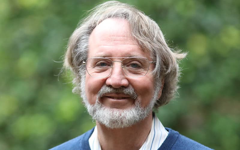 An older man with gray hair, glasses, and a beard smiles outdoors in front of blurred greenery, embodying the wisdom of elders.