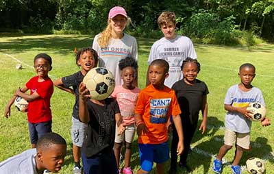A group from First Presbyterian Baton Rouge stands on grass with soccer balls, smiling at the camera, trees in the background.